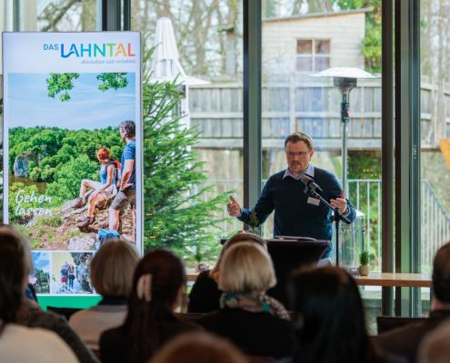 Eine Person steht an einem Rednerpult mit Mikrofon und hält eine Rede; im Hintergrund sind eine große Projektionsfolie mit Landschaftsgrafik, ein Roll-up-Banner und durch Glasfronten sichtbares Grün.