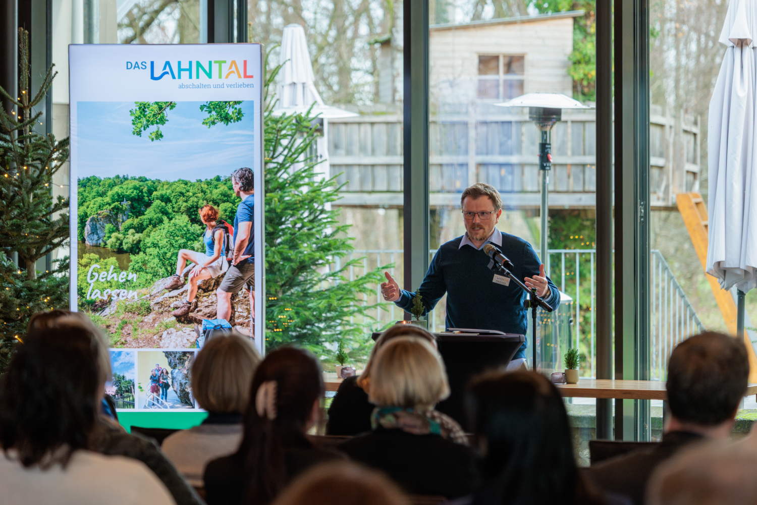 Eine Person steht an einem Rednerpult mit Mikrofon und hält eine Rede; im Hintergrund sind eine große Projektionsfolie mit Landschaftsgrafik, ein Roll-up-Banner und durch Glasfronten sichtbares Grün.
