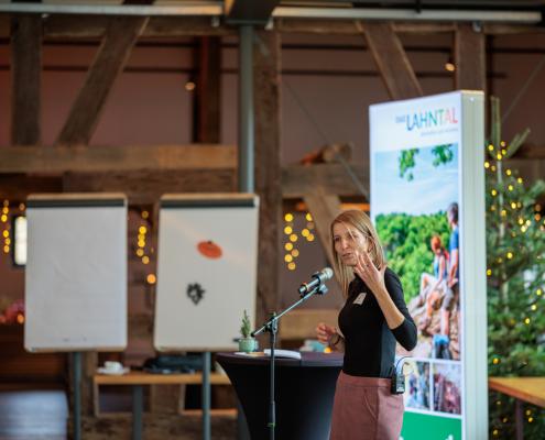 Eine Person steht an einem Rednerpult mit Mikrofon und hält eine Rede; im Hintergrund sind eine große Projektionsfolie mit Landschaftsgrafik und ein Raum mit Holzbalken