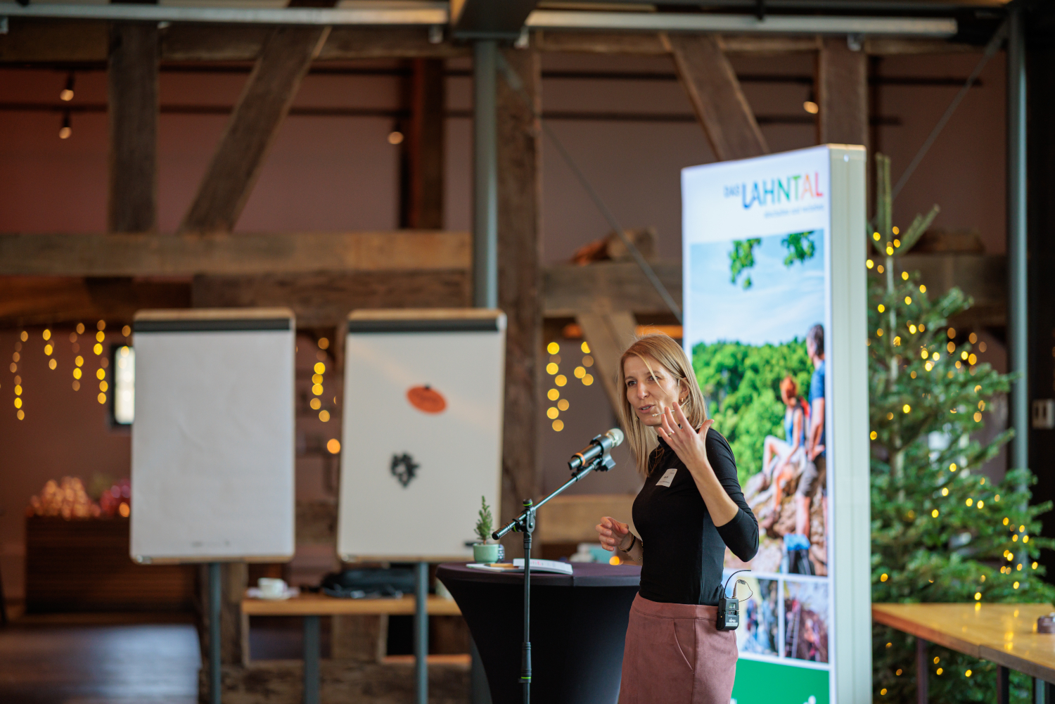 Eine Person steht an einem Rednerpult mit Mikrofon und hält eine Rede; im Hintergrund sind eine große Projektionsfolie mit Landschaftsgrafik und ein Raum mit Holzbalken