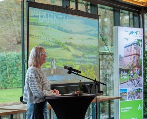 Eine Person steht an einem Rednerpult mit Mikrofon und hält eine Präsentation; im Hintergrund sind eine große Projektionsfolie mit Landschaftsgrafik, ein Roll-up-Banner und durch Glasfronten sichtbares Grün.