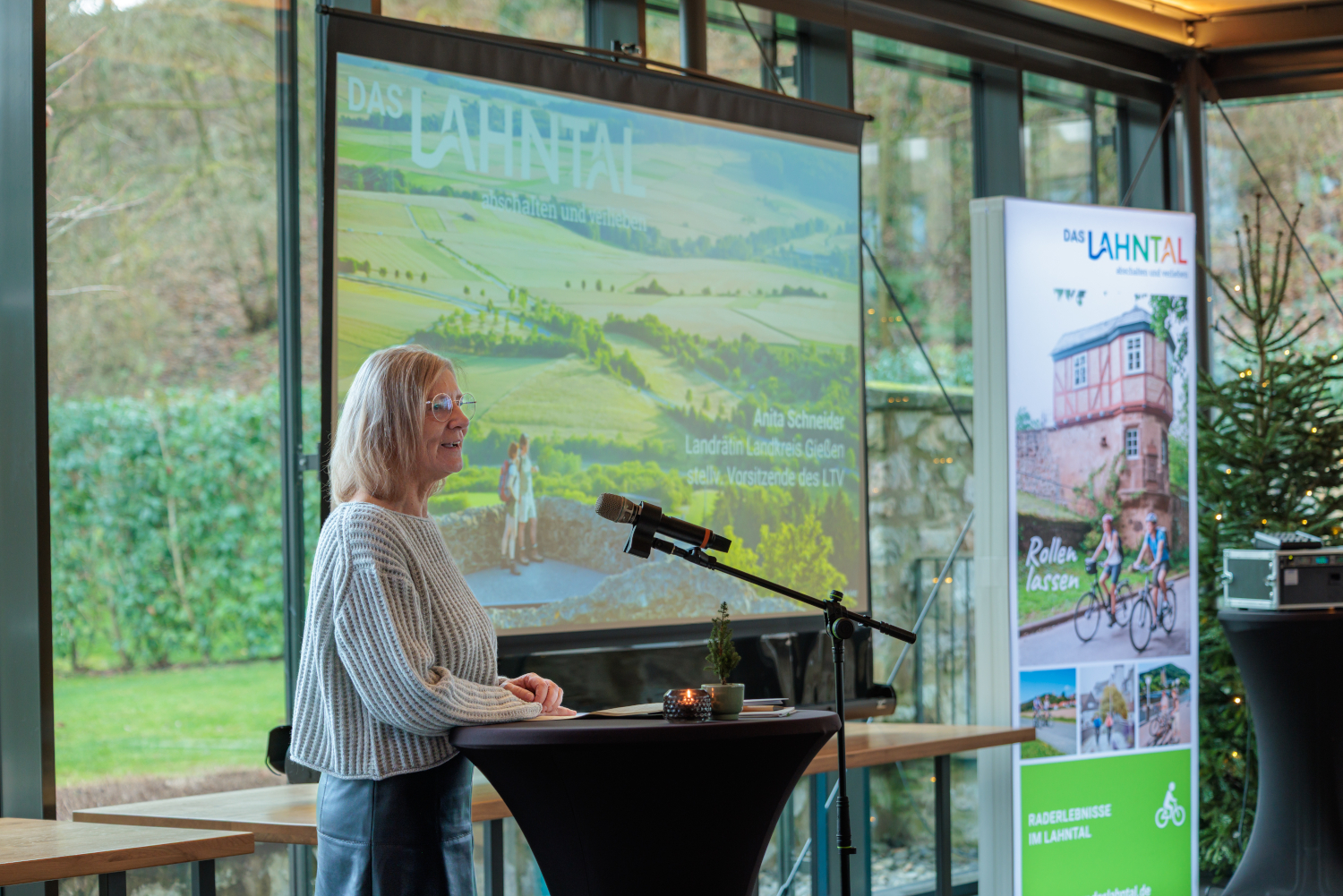 Eine Person steht an einem Rednerpult mit Mikrofon und hält eine Präsentation; im Hintergrund sind eine große Projektionsfolie mit Landschaftsgrafik, ein Roll-up-Banner und durch Glasfronten sichtbares Grün.