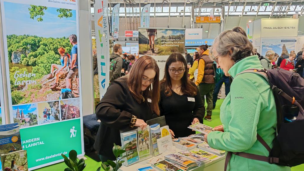 Zwei Mitarbeiterinnen informieren an einem Messestand über das Wander- und Freizeitangebot im Lahntal, während eine Besucherin Broschüren betrachtet. Im Hintergrund sind weitere Messestände und zahlreiche Besucherinnen und Besucher zu sehen.