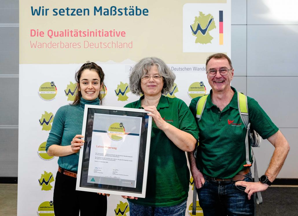Drei Personen stehen nebeneinander vor einem Banner der Qualitätsinitiative „Wanderbares Deutschland“ und halten gemeinsam eine gerahmte Urkunde in die Kamera; sie lächeln und tragen überwiegend grüne Kleidung.