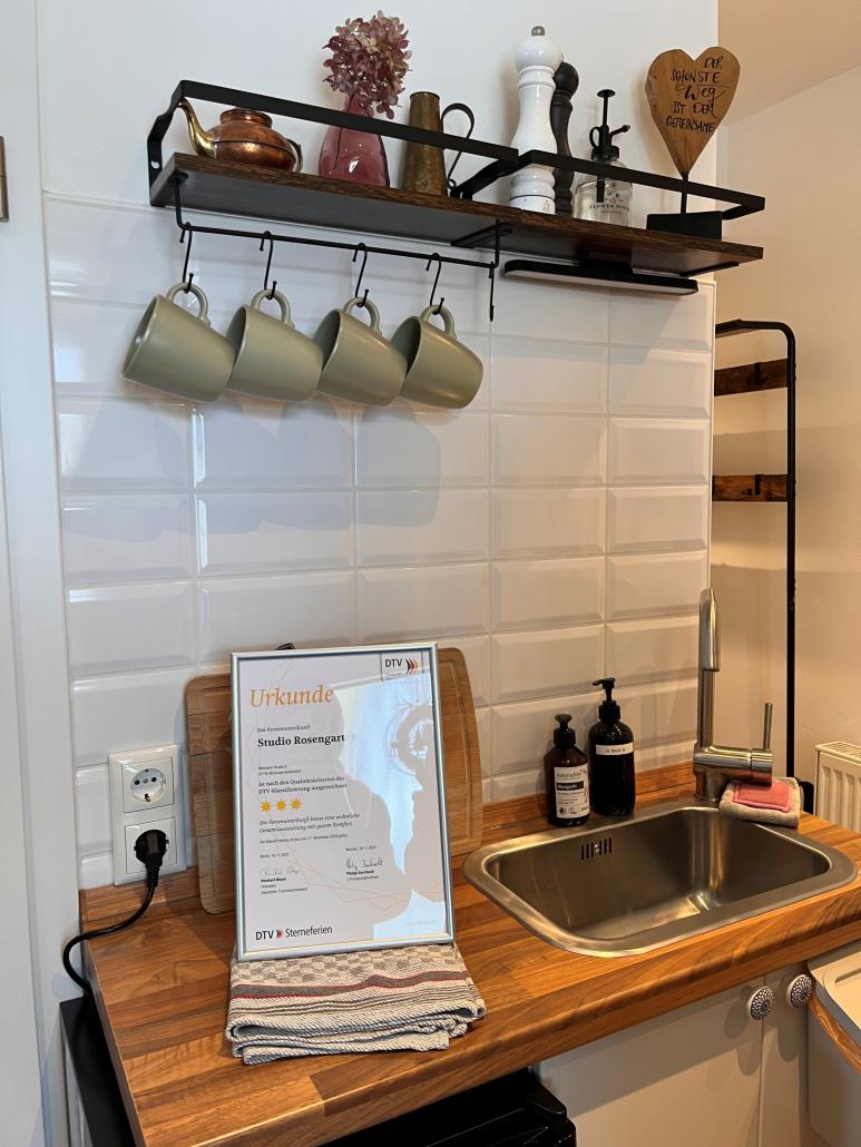 Kleine Küchenzeile mit Holzarbeitsplatte und Edelstahlspüle vor einer weißen, glänzenden Fliesenwand. Über der Spüle hängt ein schwarzes Metallregal mit Holzbrett, auf dem eine Kupferkanne, Vasen, Gewürzmühlen und ein dekoratives Holzherz stehen. Darunter hängen vier grünliche Tassen an Haken. Auf der Arbeitsfläche stehen zwei Seifenspender, ein gerahmtes Zertifikat sowie ein gefaltetes Geschirrtuch neben einer Steckdose.