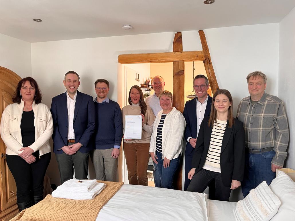 Neun erwachsene Personen stehen nebeneinander in einem hellen Raum mit sichtbaren Holzbalken und lächeln in die Kamera. In der Mitte hält eine Frau ein gerahmtes Zertifikat in den Händen. Im Vordergrund ist ein Bett mit weißer Bettwäsche zu sehen, im Hintergrund führt eine offene Tür in einen weiteren Raum.
