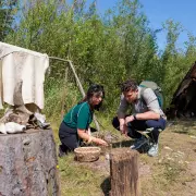 Zwei Personen knien vor einem einfachen Lager mit Zelt aus Tierhäuten und untersuchen Materialien am Boden; daneben liegen Tierknochen, ein Korb mit Fundstücken und aufgehängte Felle.