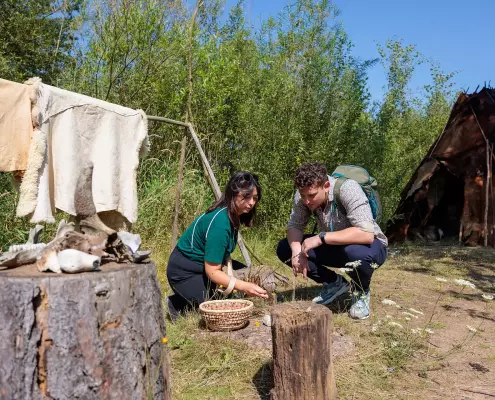 Zwei Personen knien vor einem einfachen Lager mit Zelt aus Tierhäuten und untersuchen Materialien am Boden; daneben liegen Tierknochen, ein Korb mit Fundstücken und aufgehängte Felle.