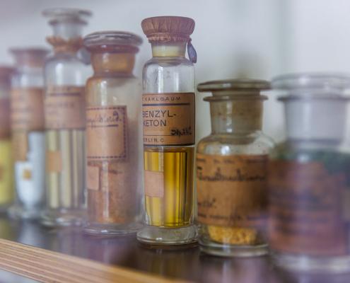 Das Foto zeigt eine Reihe antiker Apothekerfläschchen aus Glas, die ordentlich auf einem hölzernen Regalboden aufgereiht sind.Die Flaschen haben unterschiedliche Formen und Größen, sind teilweise mit gelblichen Flüssigkeiten oder Pulvern gefüllt und tragen alte, vergilbte Papieretiketten mit handschriftlichen oder gedruckten Aufschriften.