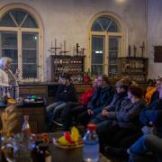 Vortrag im Liebig-Labor Eine Gruppe von Menschen sitzt in einem halbdunklen, historischen Raum mit hohen Rundbogenfenstern und hört einer älteren Frau mit grauen Haaren und heller Kleidung zu. Sie steht auf der linken Seite hinter einem Tresen, auf dem chemische Laborutensilien und Flaschen mit farbigen Flüssigkeiten aufgebaut sind. Im Hintergrund stehen alte Holzregale, die mit historischen Flaschen und Gefäßen bestückt sind, was dem Raum die Atmosphäre eines alten Labors oder einer historischen Apotheke verleiht.