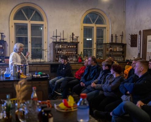 Eine Gruppe von Menschen sitzt in einem halbdunklen, historischen Raum mit hohen Rundbogenfenstern und hört einer älteren Frau mit grauen Haaren und heller Kleidung zu. Sie steht auf der linken Seite hinter einem Tresen, auf dem chemische Laborutensilien und Flaschen mit farbigen Flüssigkeiten aufgebaut sind. Im Hintergrund stehen alte Holzregale, die mit historischen Flaschen und Gefäßen bestückt sind, was dem Raum die Atmosphäre eines alten Labors oder einer historischen Apotheke verleiht.
