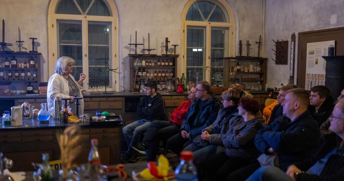 Eine Gruppe von Menschen sitzt in einem halbdunklen, historischen Raum mit hohen Rundbogenfenstern und hört einer älteren Frau mit grauen Haaren und heller Kleidung zu. Sie steht auf der linken Seite hinter einem Tresen, auf dem chemische Laborutensilien und Flaschen mit farbigen Flüssigkeiten aufgebaut sind. Im Hintergrund stehen alte Holzregale, die mit historischen Flaschen und Gefäßen bestückt sind, was dem Raum die Atmosphäre eines alten Labors oder einer historischen Apotheke verleiht.