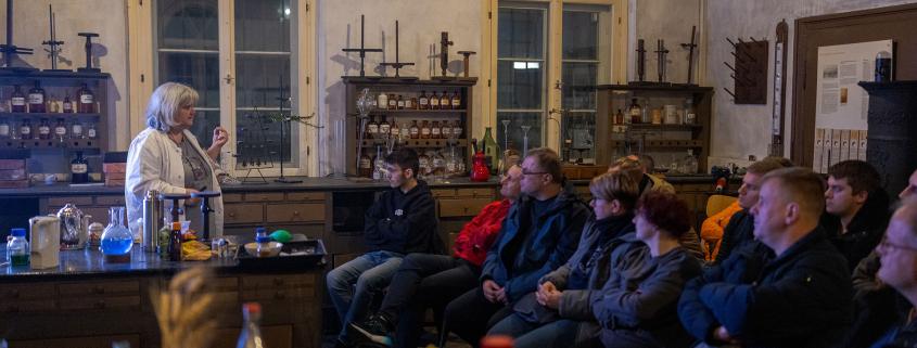 Eine Gruppe von Menschen sitzt in einem halbdunklen, historischen Raum mit hohen Rundbogenfenstern und hört einer älteren Frau mit grauen Haaren und heller Kleidung zu. Sie steht auf der linken Seite hinter einem Tresen, auf dem chemische Laborutensilien und Flaschen mit farbigen Flüssigkeiten aufgebaut sind. Im Hintergrund stehen alte Holzregale, die mit historischen Flaschen und Gefäßen bestückt sind, was dem Raum die Atmosphäre eines alten Labors oder einer historischen Apotheke verleiht.