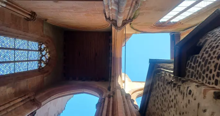 Die Perspektive zeigt den Blick steil nach oben in die unvollendete, zum Himmel offene Westfassade des Wetzlarer Doms. Die massiven Pfeiler und Spitzbögen aus rötlichem Sandstein rahmen den blauen Himmel ein, während links ein gotisches Maßwerkfenster mit blauem Muster erkennbar ist und rechts eine kontrastierende, dunklere Steinwand aufragt.
