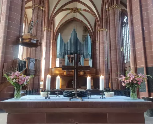Altar des Wetzlarer Doms Blick auf den altarartigen Tisch im Innenraum des Wetzlarer Doms mit einem zentralen Metallkreuz, vier brennenden Kerzen und zwei floralen Gestecken aus rosa Blumen. Im Hintergrund erhebt sich das gotische Kirchenschiff mit seinen charakteristischen Kreuzrippengewölben, rötlichen Steinsäulen und einer großen Orgel auf der Empore.