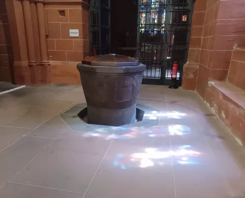 Taufbecken im Wetzlarer Dom Das historische Taufbecken aus dunklem Stein steht im Zentrum des Bildes auf einem hellen Steinboden, auf den bunte Lichtreflexe eines Buntglasfensters fallen. Im Hintergrund rahmen rötliche Sandsteinwände die Szene ein, während ein verziertes Metallgitter den Blick in eine dahinterliegende Kapelle freigibt.