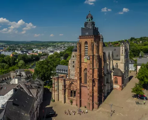 Der Wetzlarer Dom Die Luftaufnahme zeigt den monumentalen Wetzlarer Dom aus der Vogelperspektive, umgeben von historischen Dächern und einer grünen Stadtlandschaft. Im Zentrum steht der unvollendete, rötlich-braune Südturm, hinter dem das hellere Kirchenschiff aufragt.