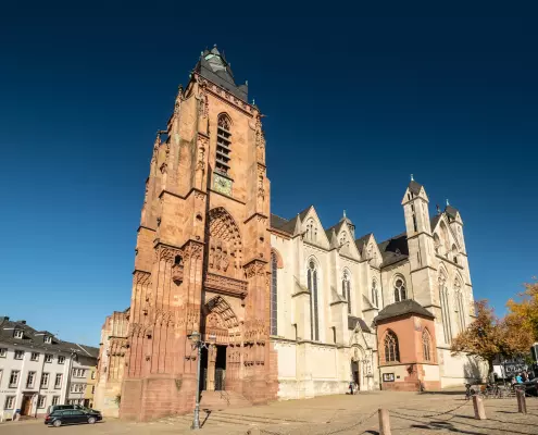 Der Wetzaler Dom Ein großer, rötlich-brauner Sandsteinturm dominiert die linke Seite der Kirchenfassade, die durch gotische Bögen und Skulpturen über dem Portal verziert ist. Rechts schließt sich das hellere Hauptschiff an, das mit spitz zulaufenden Fenstern und Giebeln gestaltet ist.