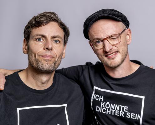 Simon & Jan, ein deutsches Liedermacher-Duo Ein Porträtfoto von Simon & Jan, einem deutschen Liedermacher-Duo. Die beiden Männer stehen eng nebeneinander und tragen schwarze Langarmshirts mit einem weißen quadratischen Aufdruck.Links ist Simon Eickhoff zu sehen, der mit einem neutralen, leicht lächelnden Gesichtsausdruck direkt in die Kamera blickt. Er hat braunes, leicht gewelltes Haar und einen Dreitagebart.Rechts steht Jan Traphan, der eine dunkle Schiebermütze und eine Brille mit rötlichem Gestell trägt. Er zwinkert mit dem linken Auge und grinst verschmitzt.Auf Jans Shirt ist innerhalb des weißen Quadrats der Text „ICH KÖNNTE DICHTER SEIN“ in weißen Großbuchstaben zu lesen – ein humorvolles Wortspiel, das sowohl auf poetisches Talent als auch auf Alkoholkonsum anspielen kann. Der Hintergrund des Bildes ist schlicht und in einem hellen Grauton gehalten.