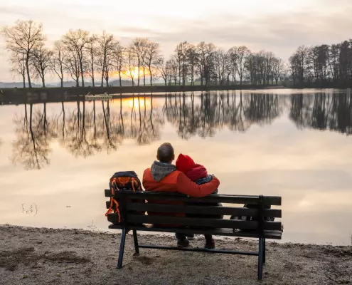 Ein Mann und eine Frau mit einer roten Mütze sitzen Arm in Arm auf einer Holzbank am Ufer eines stillen Sees und blicken auf den Sonnenuntergang. Am gegenüberliegenden Ufer spiegelt sich eine Reihe von kahlen Bäumen perfekt auf der glatten Wasseroberfläche, während der Himmel in sanften Orange- und Grautönen leuchtet. Links auf der Bank steht ein Rucksack, und der Vordergrund zeigt einen kiesigen Boden mit spärlichem Gras.