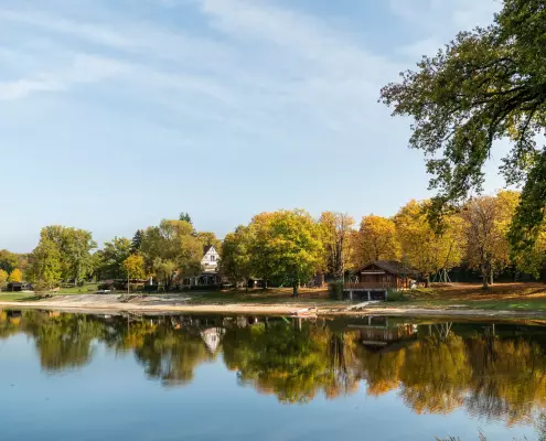 Ein idyllischer See an einem sonnigen Herbsttag, an dessen bewaldetem Ufer sich Bäume mit goldgelbem Laub und ein Holzhaus im ruhigen Wasser spiegeln.