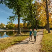 Ein Mann und eine Frau wandern an einem sonnigen Herbsttag auf einem geschotterten Weg entlang eines Sees. Rechts des Weges reihen sich große Bäume mit grünem und goldgelb gefärbtem Laub aneinander. Auf der linken Seite spiegelt sich das gegenüberliegende Ufer mit weiteren Bäumen und vereinzelten Gebäuden im ruhigen Wasser des Sees.
