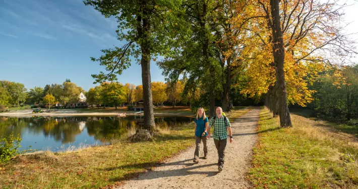 Ein Mann und eine Frau wandern an einem sonnigen Herbsttag auf einem geschotterten Weg entlang eines Sees. Rechts des Weges reihen sich große Bäume mit grünem und goldgelb gefärbtem Laub aneinander. Auf der linken Seite spiegelt sich das gegenüberliegende Ufer mit weiteren Bäumen und vereinzelten Gebäuden im ruhigen Wasser des Sees.