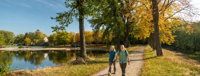 Ein Mann und eine Frau wandern an einem sonnigen Herbsttag auf einem geschotterten Weg entlang eines Sees. Rechts des Weges reihen sich große Bäume mit grünem und goldgelb gefärbtem Laub aneinander. Auf der linken Seite spiegelt sich das gegenüberliegende Ufer mit weiteren Bäumen und vereinzelten Gebäuden im ruhigen Wasser des Sees.