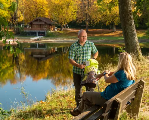 Ein Mann im grün karierten Hemd steht mit einer Wanderkarte und einem hellgrünen Rucksack an einem herbstlichen Seeufer und spricht mit einer blonden Frau, die auf einer Holzbank sitzt. Im Hintergrund spiegelt sich ein Holzhaus im ruhigen Wasser, umgeben von Bäumen mit gelb-orange gefärbtem Laub.