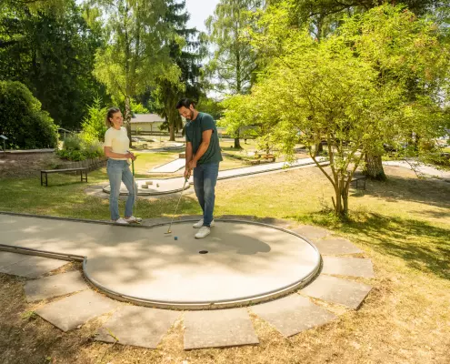 Ein Mann mit Bart und kurzen dunklen Haaren steht leicht nach vorne gebeugt auf einer Minigolfbahn, hält einen Minigolfschläger mit beiden Händen und zielt auf einen kleinen blauen Ball, während er Jeans und ein dunkelgrünes T-Shirt trägt.
