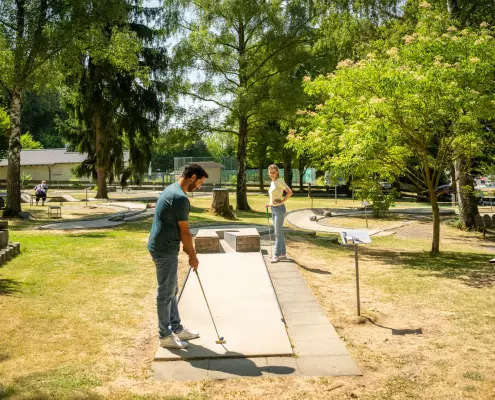 in Mann und eine Frau spielen an einem sonnigen Tag Minigolf auf einer von Bäumen umgebenen Anlage im Freien. Der Mann im Vordergrund trägt ein dunkelgrünes T-Shirt und Jeans, steht in gebückter Haltung auf der Betonbahn und setzt gerade mit dem Minigolfschläger zum Schlag an. Die Frau steht etwas weiter hinten im Hintergrund, trägt ein helles T-Shirt sowie Jeans, hält ebenfalls einen Schläger und schaut lächelnd in Richtung des Mannes.