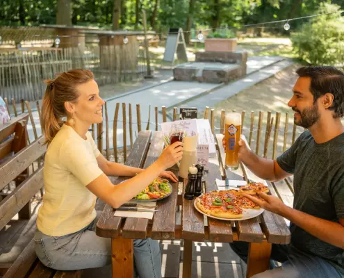 Ein Mann und eine Frau sitzen lächelnd an einem rustikalen Holztisch im Außenbereich eines Lokals und stoßen mit ihren Getränken an. Vor ihnen liegen eine Pizza und ein Salat auf dem Tisch, während im Hintergrund eine Minigolfanlage im Grünen zu sehen ist.