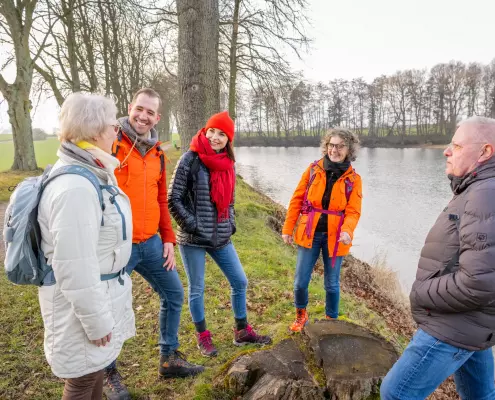 Eine fünfköpfige Gruppe verschiedener Generationen steht gut gelaunt an einem Uferweg neben einem See und unterhält sich. Die Wanderer tragen wetterfeste Outdoorkleidung wie Daunen- und Regenjacken in auffälligem Orange, Schwarz, Grau und Weiß sowie Rucksäcke. Im Vordergrund befindet sich ein alter Baumstumpf, während im Hintergrund eine Allee aus kahlen Bäumen und eine weite Wiese unter leicht bewölktem Himmel zu sehen sind.
