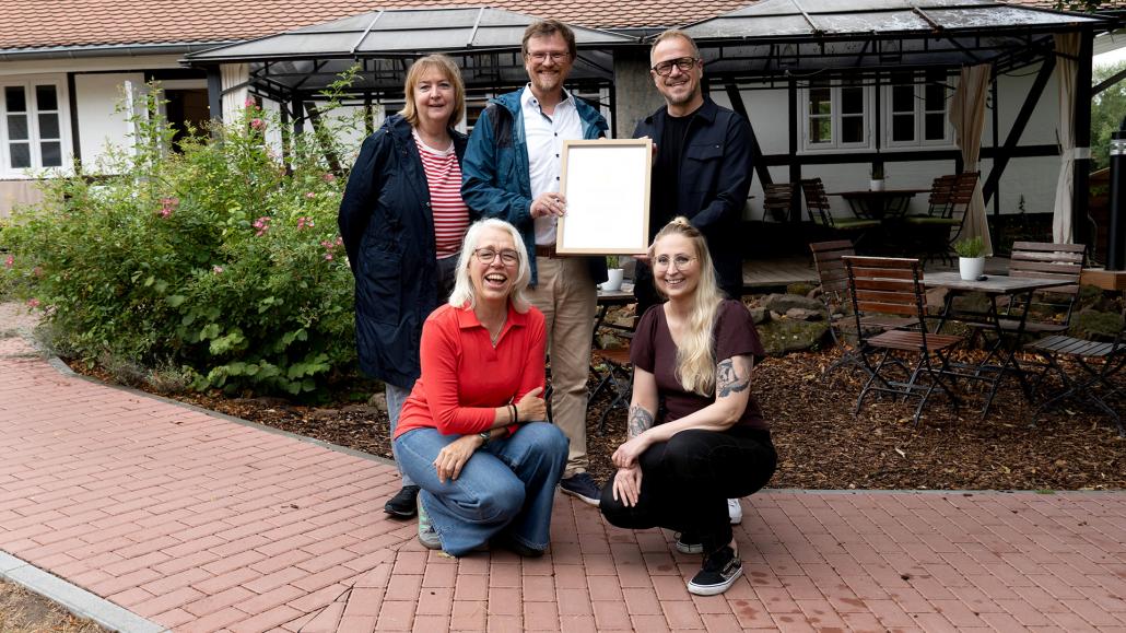 Vertreter des Lahntal Tourismus Verbands übergeben lächelnd eine gerahmte Urkunde vor einem Fachwerkhaus an das Team der Fleckenbühler.