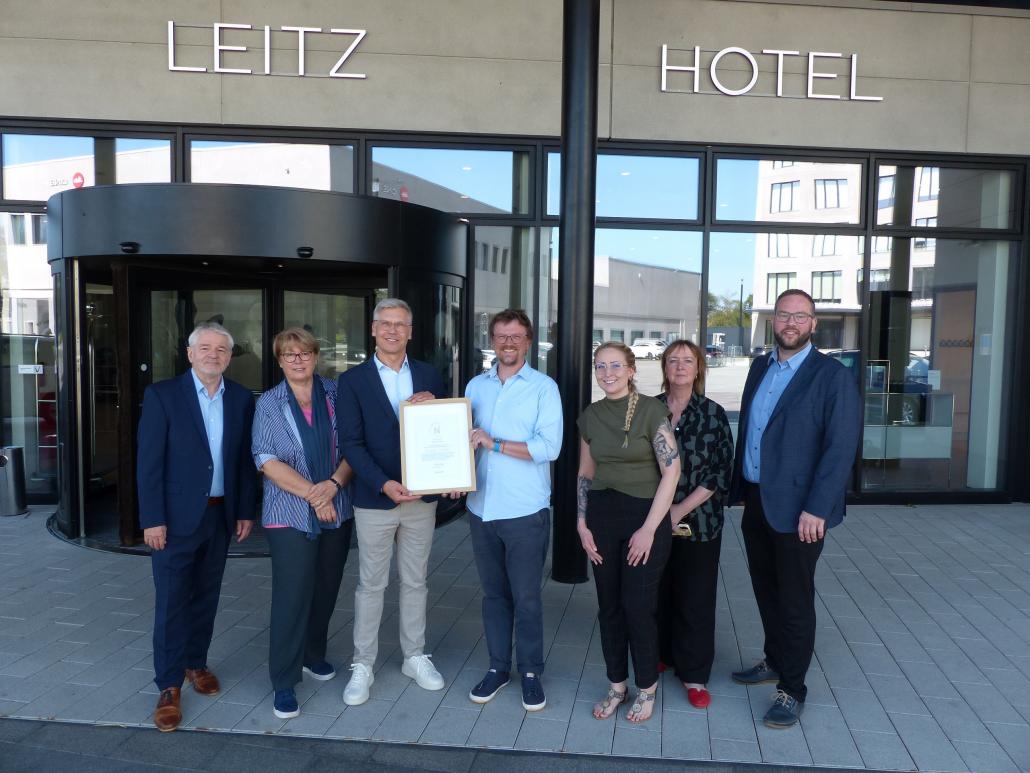 Das Bild zeigt eine feierliche Übergabe einer gerahmten Urkunde vor dem Eingang des Ernst Leitz Hotels in Wetzlar. Eine Gruppe von sieben Personen steht lächelnd nebeneinander vor der markanten Glasfassade und der modernen Drehtür des Hotelgebäudes, über dem in silbernen Lettern der Schriftzug „LEITZ HOTEL“ zu sehen ist.