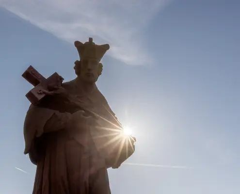 Johannes-Nepomuk-Statue in Amöneburg Silhouette einer Heiligenstatue mit Kruzifix vor blauem Himmel und strahlender Sonne.