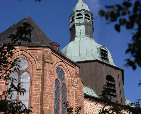 Stiftskirche St. Johannes der Täufer in Amöneburg Blick durch grünes Laub auf den markanten barocken Kirchturm und die neugotische Fassade der Stiftskirche St. Johannes der Täufer in Amöneburg vor blauem Himmel.