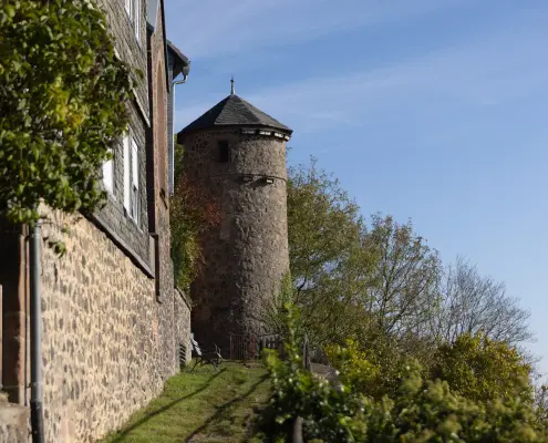 Ansicht von Amöneburg Ein runder, historischer Steinturm mit spitzem Dach steht hinter einer alten Natursteinmauer an einem bewachsenen Hang unter blauem Himmel.