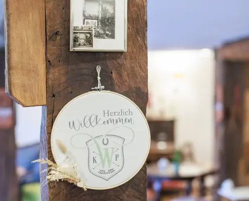 Café-Dekoration Runder Stickrahmen mit der Aufschrift „Herzlich Willkommen“ und einem Familienwappen mit den Buchstaben K, W und P, geschmückt mit Trockenblumen.