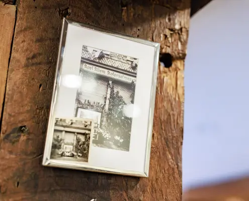 Café-Dekoration Ein schlichter Silberrahmen hängt an einem Holzbalken und zeigt historische Schwarz-Weiß-Aufnahmen, darunter eine mit dem Schriftzug „Karl Kliem Kolonialwaren“.