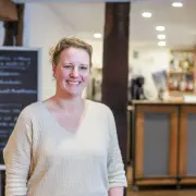 Eine lächelnde Frau steht im Vordergrund eines gemütlich eingerichteten Gastraums oder Restaurants mit sichtbaren Holzbalken, einer Schiefertafel im Hintergrund und einer Theke.