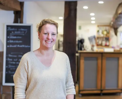 Eine lächelnde Frau steht im Vordergrund eines gemütlich eingerichteten Gastraums oder Restaurants mit sichtbaren Holzbalken, einer Schiefertafel im Hintergrund und einer Theke.