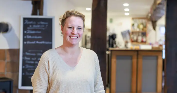Eine lächelnde Frau steht im Vordergrund eines gemütlich eingerichteten Gastraums oder Restaurants mit sichtbaren Holzbalken, einer Schiefertafel im Hintergrund und einer Theke.
