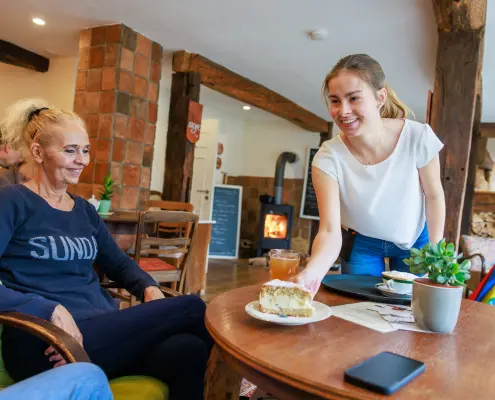 Kuchenpause im Café Die Person auf dem Foto sitzt in einem gemütlichen Café an einem Holztisch, während eine Kellnerin ein Stück Torte serviert.