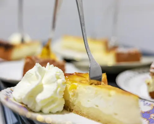 Ein Stück Käsekuchen mit Sahne Ein Stück Käsekuchen mit einer Kuchengabel im oberen Bereich und einem großen Klecks Schlagsahne auf einem gemusterten Teller.