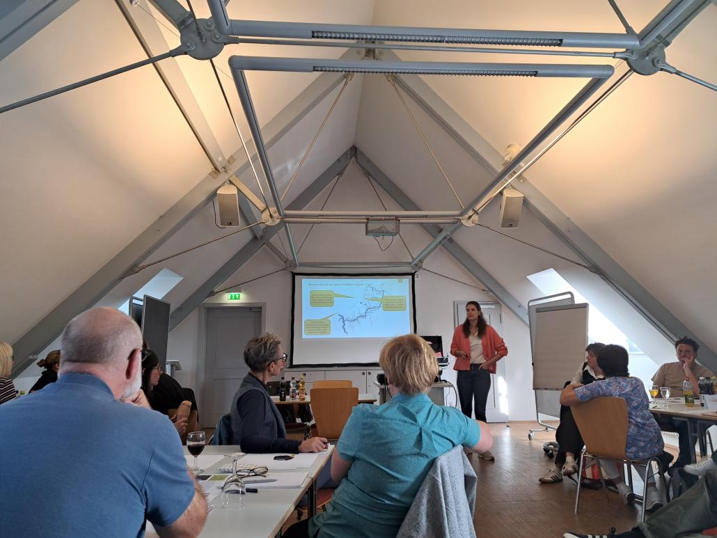 Workshop mit mehreren Teilnehmenden in einem Seminarraum. Die Gruppe sitzt an Tischen und arbeitet gemeinsam, während vorne eine Präsentation auf einer Leinwand gezeigt wird. Eine Person steht neben der Präsentation und moderiert den Workshop.