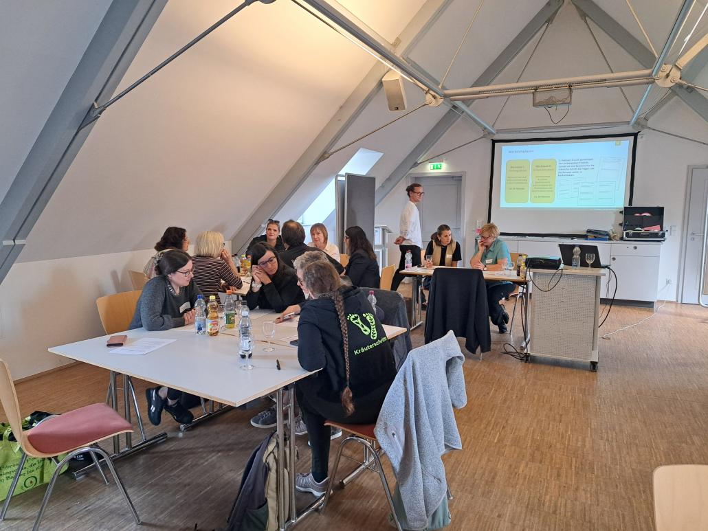 Workshop mit mehreren Teilnehmenden in einem Seminarraum. Die Gruppe sitzt an Tischen und arbeitet gemeinsam, während vorne eine Präsentation auf einer Leinwand gezeigt wird.