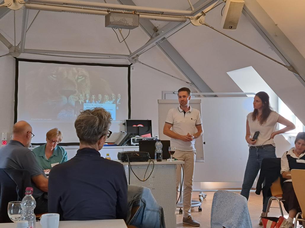 Eine Präsentation in einem hellen Seminarraum unter einem Dachgeschoss. Im Hintergrund stehen zwei Referenten (ein Mann im weißen Poloshirt und eine Frau in Jeans) vor einer großen Leinwand, auf der eine Folie mit einem Löwenmotiv projiziert wird. Im Vordergrund sitzt das Publikum an Tischen und folgt dem Vortrag.