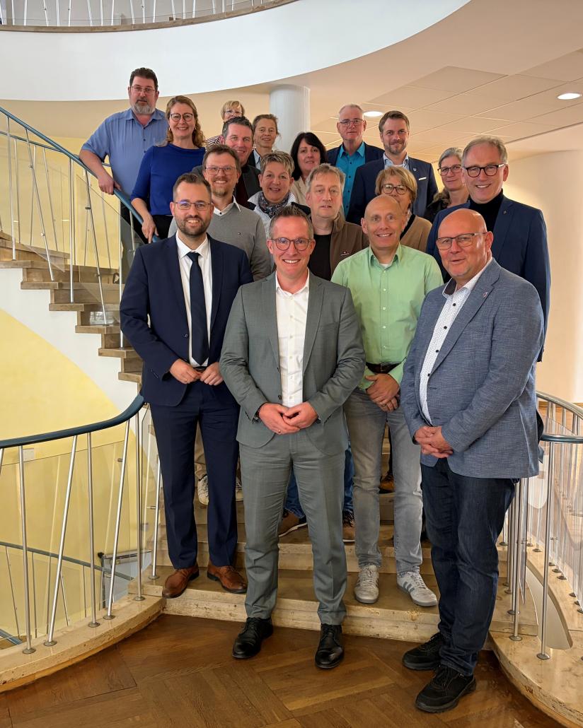 Das Bild zeigt eine Gruppe von 18 Personen, die sich für ein Gemeinschaftsfoto auf den Stufen einer großen, geschwungenen Wendeltreppe im Innenbereich aufgestellt hat.